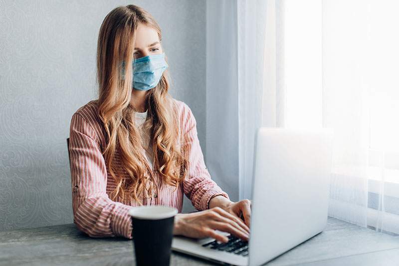 A young businesswoman works from home, sitting at a table with a laptop, wearing a protective mask. The concept of quarantine, coronavirus